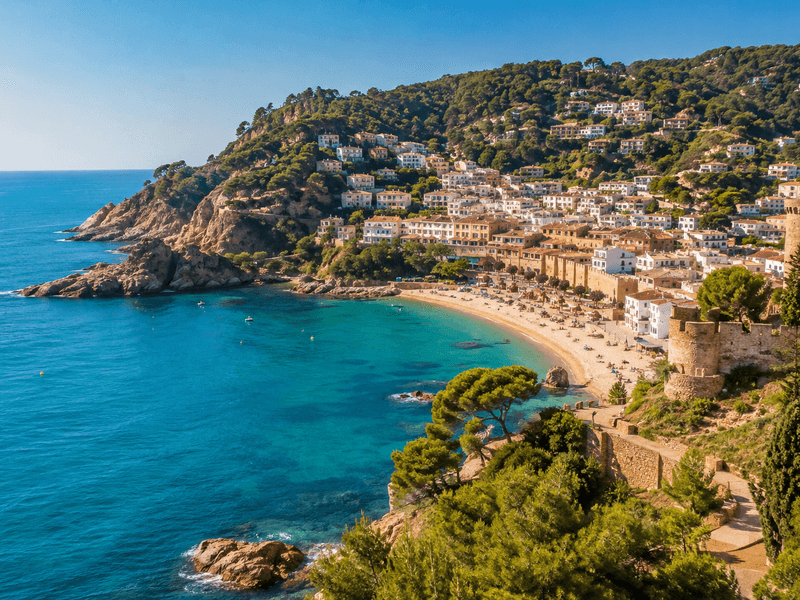 Vista panorámica de una localidad costera de la Costa Brava con viviendas en la ladera, playa, mar Mediterráneo y torres de piedra